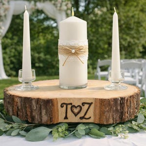 May include: A rustic wedding centerpiece featuring a wooden slab with a carved "T♥Z" design. It holds a large pillar candle with lace and twine, flanked by two taper candles in glass holders. Greenery surrounds the base.