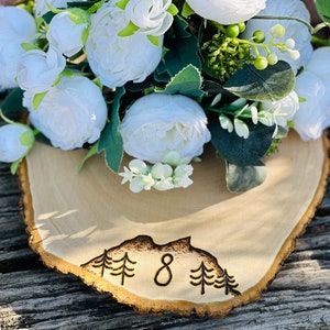 May include: A wooden slice with a burnt design of a mountain range and the number 8. A bouquet of white flowers sits on top of the slice.