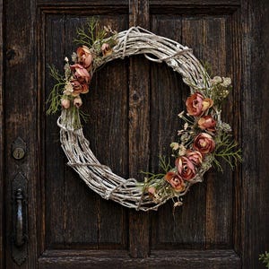 May include: A circular wreath made of woven, light-colored branches, adorned with faux peach-colored roses and green sprigs. The wreath hangs on a dark brown wooden door, creating a rustic and natural aesthetic.