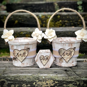 May include: Two white-washed wooden flower girl baskets with rope handles and floral accents. Each basket features a heart-shaped wood cutout with the initials "A+M" and an arrow. A matching hexagon-shaped box is also present.