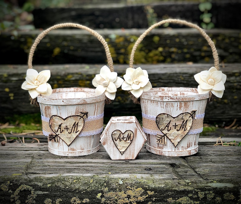 May include: Two white-washed wooden flower girl baskets with rope handles and floral accents. Each basket features a heart-shaped wood cutout with the initials "A+M" and an arrow. A matching hexagon-shaped box is also present.