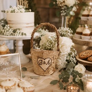 May include: A rustic birch bark basket with a woven handle, decorated with a heart-shaped emblem featuring the initials "B&A". The basket is surrounded by white roses and greenery, part of a wedding reception display.