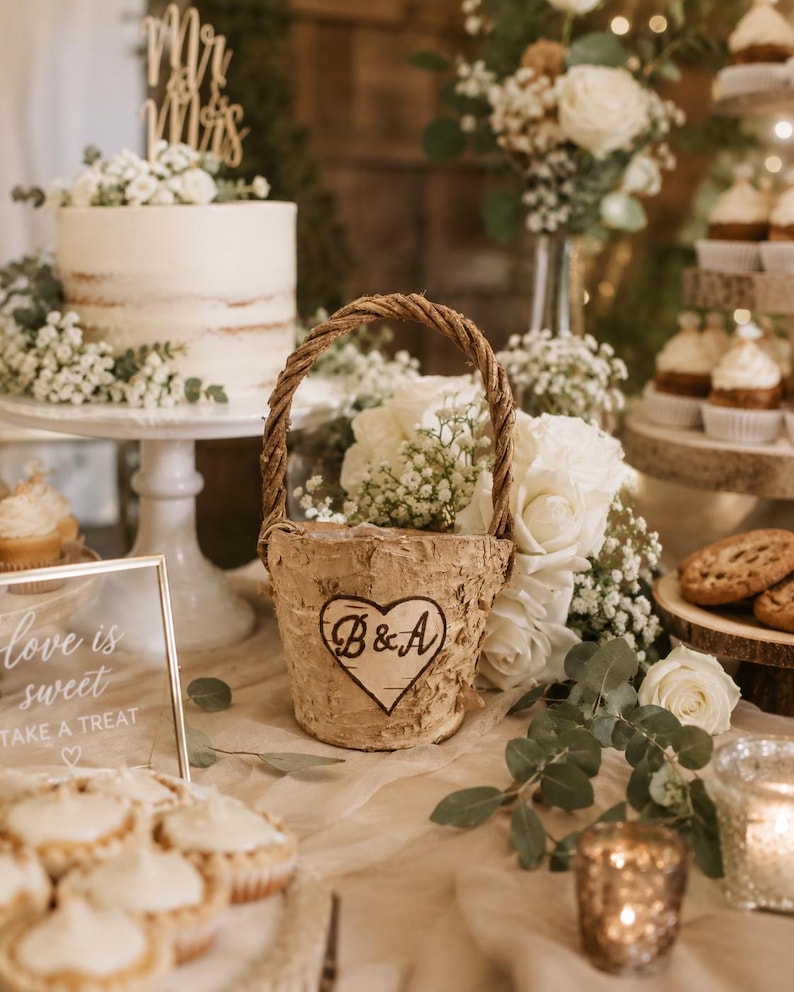 May include: A rustic birch bark basket with a woven handle, decorated with a heart-shaped emblem featuring the initials "B&A". The basket is surrounded by white roses and greenery, part of a wedding reception display.