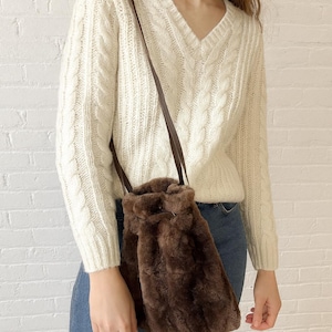 May include: A brown faux fur bucket bag with a drawstring closure and a long brown strap. The bag is worn over the shoulder. The person is wearing a cream-colored cable knit sweater and blue jeans.