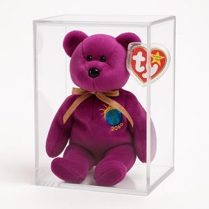 Puede incluir: Un oso de peluche morado con un lazo de cinta dorada y un emblema de globo, exhibido en una caja transparente. El oso tiene una etiqueta en forma de corazón. El emblema en el oso dice "2060".