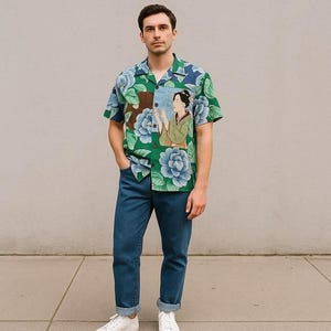 May include: A short-sleeved shirt with a green base and large blue floral patterns. The shirt features an illustration of a woman in traditional dress. The person is wearing blue jeans and white trainers.