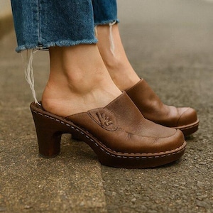 Pode incluir: Tamancos de couro castanho com salto alto e bico arredondado. Os tamancos têm um design slip-on e um emblema decorativo na parte superior. A sola da plataforma é costurada com uma linha contrastante.