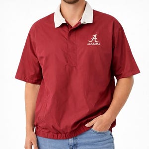 May include: A crimson short-sleeve pullover shirt with a white collar. The shirt features the word "ALABAMA" under a stylized "A" logo. The shirt has a drawstring hem and a buttoned placket.