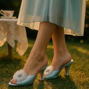 Puede incluir: Mules de satén azul claro con un adorno floral. Los tacones abiertos tienen un tacón redondeado y se usan sobre hierba. Un vestido azul claro es visible en el fondo, junto con una pequeña mesa y una taza de té.