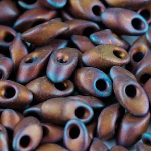 May include: A close-up of a pile of small, brown and blue glass beads. The beads are oval shaped and have a matte finish.
