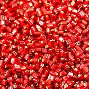 May include: A close-up image of a large quantity of small, red, glass seed beads. The beads are all uniform in size and shape, and they have a slightly iridescent finish.