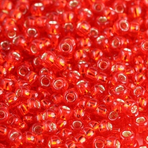May include: A close-up view of a large quantity of small, red glass beads. The beads are round with a small hole in the center. The beads have a shiny, metallic finish.