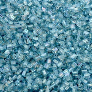May include: A close-up view of a large quantity of small, light blue glass beads. The beads are iridescent and have a slightly rounded, rectangular shape.