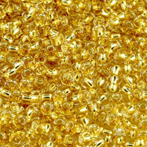 May include: Close-up of a pile of small, golden-yellow glass beads. The beads are cylindrical with rounded edges and a hole running through the center. The beads are densely packed together, creating a shimmering, textured surface. Ideal for jewelry making and crafts.