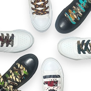 Puede incluir: Cinco pares de zapatillas blancas con cordones de diferentes patrones. Los cordones son de varios colores y patrones, incluyendo negro, rojo, verde, azul y amarillo.