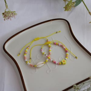 Könnte beinhalten: Zwei bunte Perlenarmbänder mit gelben Schnüren. Ein Armband hat einen weißen Muschel-Anhänger und das andere einen rosa-gelben Blumen-Anhänger.