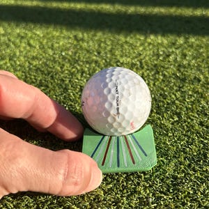 May include: A white golf ball with a dimpled surface and the text NXT TOUR S printed on it, resting on a light green plastic stand featuring a series of thin, radiating lines in dark green, red, and silver colors across its curved front surface.