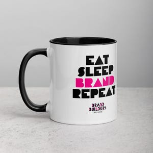 May include: White ceramic mug with a black interior and handle. The mug features the text "EAT SLEEP BRAND REPEAT" in black and pink lettering. The words "BRAND BUILDERS ALLIANCE" are printed at the bottom in a smaller font.