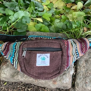 May include: A fanny pack featuring a brown woven front pocket with a white label that reads HIMALAYAN HEMP THC FREE SEEDLESS HEMP and a green leaf icon. The bag has a multi-colored striped pattern in shades of teal, maroon, yellow, and black.