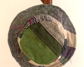Może przedstawiać: Czapka typu bucket hat wykonana z różnych kawałków materiału, z szerokim rondem w szaro-czarny wzór w jodełkę. Główka czapki prezentuje mieszankę faktur i kolorów, w tym wyrazisty jasnozielony panel, beż oraz odcienie niebieskiego.