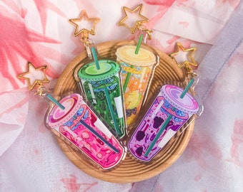 May include: Four colorful acrylic drink keychains with gold star-shaped clasps. The drinks are pink, green, orange, and purple, each with a straw. They are arranged on a small wooden tray, set against a floral background.