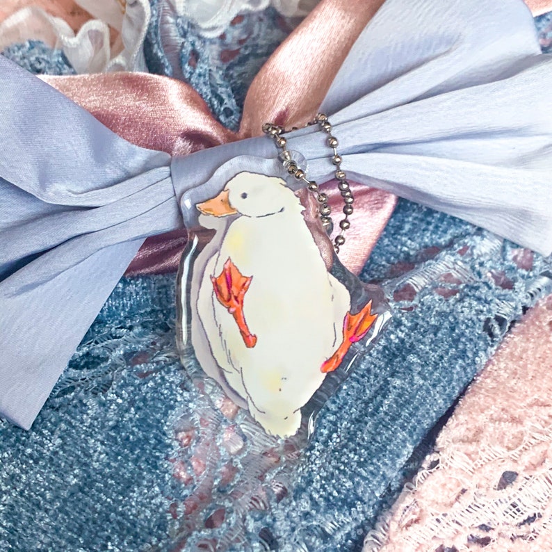 May include: A clear acrylic charm featuring a whimsical illustration of a white duck with orange feet and beak. The charm hangs from a silver ball chain, set against a backdrop of blue and pink ribbons and lace.