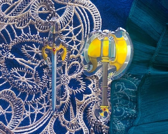 May include: Two clear acrylic charms featuring a sword and an axe. The sword is gold with a red jewel and the axe is gold with a black handle. The charms are hanging from a silver chain against a blue and green fabric background.