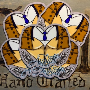 Könnte beinhalten: Ein Stapel herzförmiger Aufkleber mit einem Cartoon-Design des Unterkörpers einer Person in einem Anzug. Die Aufkleber haben einen hellbraunen Körper, eine braune Jacke, ein weißes Hemd und eine blaue Krawatte. Das Wort "Assbutt" ist in Schreibschrift mit Engelsflügeln geschrieben.