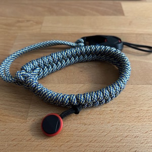 May include: A blue and yellow braided wrist strap with a black and red circular attachment. The strap is made of a thick, durable rope-like material. A black strap with a loop is attached to the wrist strap.