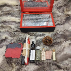 May include: A red and black wooden box with a mirrored interior sits atop a faux fur surface. Various items are arranged around it, including candles, small bottles, and a dried seed pod. One bottle has a label that reads "PROTECTION & REVERSAL".