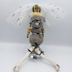 May include: A handcrafted doll-like figure with a unique design. It features a headpiece with yellow and black accents, a mesh body, and bone-like legs. The figure is adorned with decorative elements and a white, veil-like structure.
