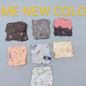 May include: The image displays six textured, square-shaped paper samples in various colors, including brown, gray, pink, peach, white, and a speckled mix. The text "SOME NEW COLORS" is in yellow above the samples.