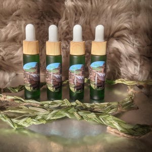 Może przedstawiać: Cztery małe zielone butelki szklane z zakraplaczami i drewnianymi korkami. Butelki są oznaczone napisem "Sage" i mają naturalny design. Butelki stoją na jasnobrązowej powierzchni z plecionym zielonym roślinem.