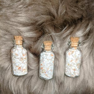 May include: Three small glass bottles with cork stoppers filled with white and orange iridescent glitter. The bottles are sitting on a brown fuzzy surface.