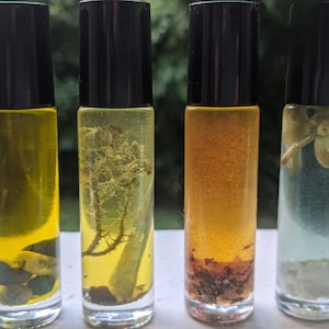 Puede incluir: Cuatro botellas de vidrio con rodillo y tapas negras. Las botellas contienen líquidos de diferentes colores y diversos elementos botánicos. Las botellas están dispuestas en fila sobre una superficie blanca.