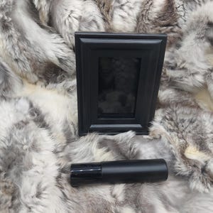 May include: A black rectangular picture frame and a black cylindrical object are displayed on a textured, gray and white surface. The frame is upright, and the cylindrical object is horizontal.