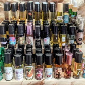 May include: A wooden shelf display with multiple small glass bottles containing essential oils. The bottles have black roll-on tops and colorful labels with various designs and text, including "Healing", "Fire", "7", and "Forbidden Royalty".