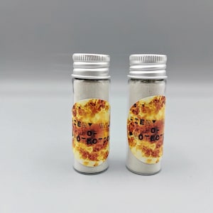Puede incluir: Dos pequeños frascos de vidrio con tapas plateadas, llenos de un polvo de color claro. Cada frasco tiene una etiqueta con el texto "Fiery Way of Protection" en una fuente estilizada, sobre un fondo naranja y amarillo llameante.