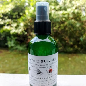May include: A green glass spray bottle with a black cap and a clear plastic top. The label reads "DON'T BUG ME" with illustrations of insects. The bottle contains a liquid and is from "ENCHANTED EMPORIUM".