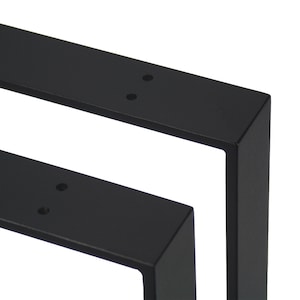 Op de afbeelding: Close-up van twee zwarte metalen tafelpoten. Elke poot heeft een rechthoekige vorm met een haakse hoek en voorgeboorde gaten voor montage. De poten zijn ontworpen voor meubelconstructie.