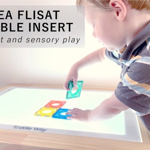 Puede incluir: Un niño juega con azulejos magnéticos de colores en un inserto de mesa iluminado para la mesa IKEA FLISAT. El inserto de mesa está etiquetado como "Toddler Way".