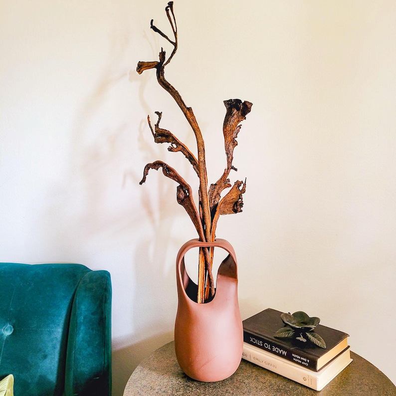 Op de afbeelding: Een terracotta-kleurige vaas met een uniek handvatontwerp, gevuld met gedroogde takken. De vaas staat op een ronde tafel naast een stapel boeken en een decoratieve metalen bloem. Op de achtergrond is een turquoise fauteuil te zien.