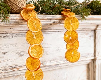 May include: A festive garland of dried orange slices hangs from a rustic mantelpiece, adorned with evergreen sprigs and twine. The warm orange hues of the citrus contrast with the green of the pine, creating a cozy, natural holiday decoration.