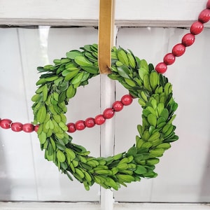 May include: A fresh green boxwood wreath with a gold ribbon for hanging. A strand of red wooden beads is draped across the wreath. The wreath is hanging in front of a white window.