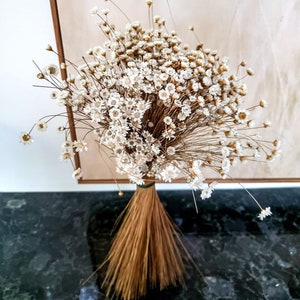 May include: A bouquet of dried white flowers with small daisy-like blooms. The stems are bundled together with a natural fiber and tied with green twine. The bouquet is set against a neutral background.