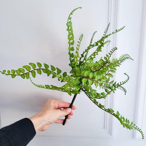 May include: A green artificial fern with small, oval-shaped leaves and a dark stem. The fern has multiple stems that curve and arch, creating a natural, flowing appearance. The leaves are a vibrant green color, and the stem is dark brown.