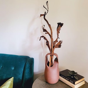 Op de afbeelding: Een terracotta vaas met een uniek handvat, gevuld met gedroogde takken en bruine bladeren. De vaas staat op een ronde, grijze tafel naast boeken en een metalen bloemversiering. Op de achtergrond is een turquoise bank te zien.