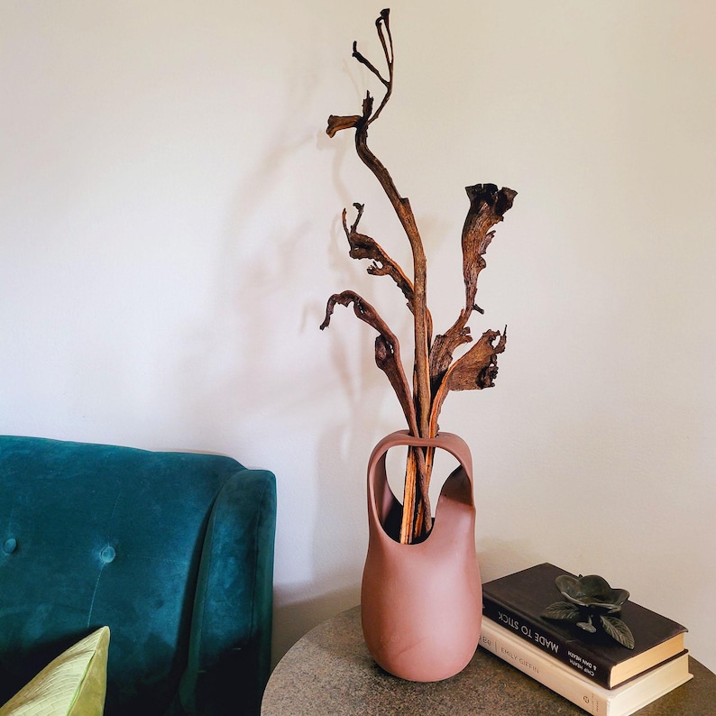 Op de afbeelding: Een terracotta vaas met een uniek handvat, gevuld met gedroogde takken en bruine bladeren. De vaas staat op een ronde, grijze tafel naast boeken en een metalen bloemversiering. Op de achtergrond is een turquoise bank te zien.