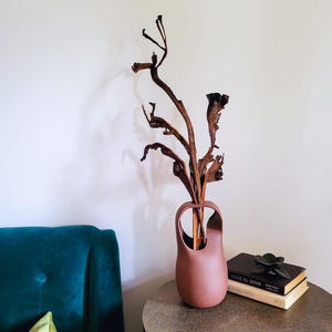 Op de afbeelding: Een hoge, terracotta-kleurige vaas met een uniek handvatontwerp, met gedroogde takken. De vaas staat op een ronde, metalen bijzettafel naast een stapel boeken en een decoratieve bloem. De achtergrond is een witte muur en een blauwgroene stoel.