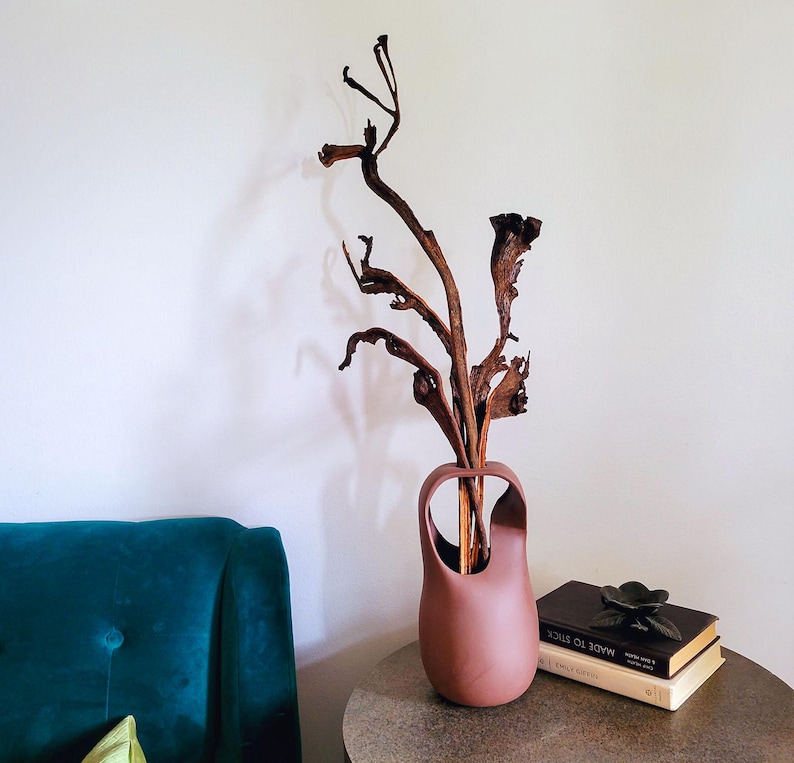 Op de afbeelding: Een hoge, terracotta-kleurige vaas met een uniek handvatontwerp, met gedroogde takken. De vaas staat op een ronde, metalen bijzettafel naast een stapel boeken en een decoratieve bloem. De achtergrond is een witte muur en een blauwgroene stoel.